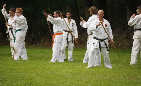 Karateka's met oranje en groene band oefenen met de jo bij de bosrand