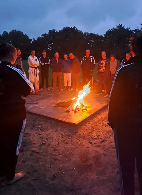 Deelnemers staan 's avonds samen rond het kampvuur