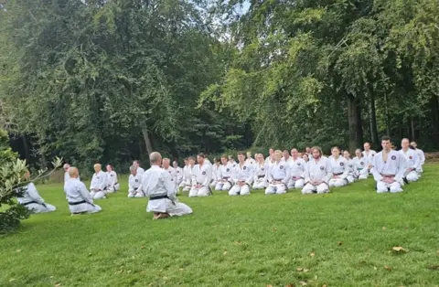 Groep zit in seiza op het gras terwijl de sensei uitleg geeft